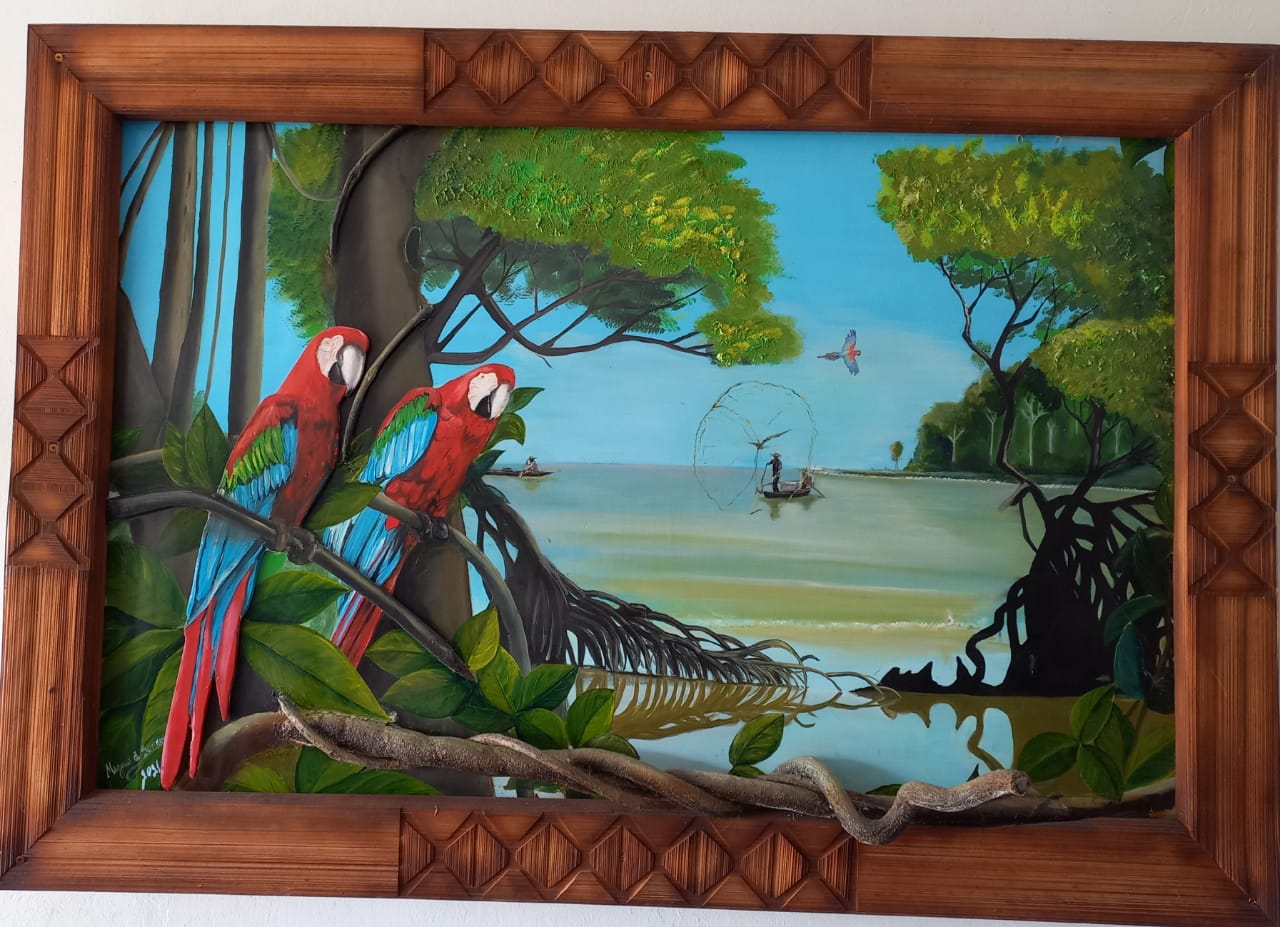 Pintura em relevo de duas araras-vermelhas pousadas em galhos de árvores que saem da moldura. Elas estão em primeiro plano, à esquerda. Ao fundo, uma paisagem amazônica com um rio calmo, vegetação de mangue e um pescador em uma pequena canoa lançando uma rede circular (tarrafa). O céu é azul claro.